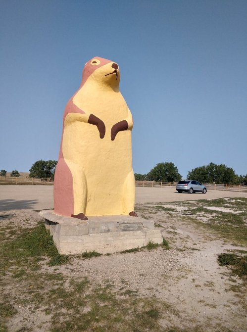 A large, colorful statue of a prairie dog stands in an open area with a car parked nearby and trees in the background.