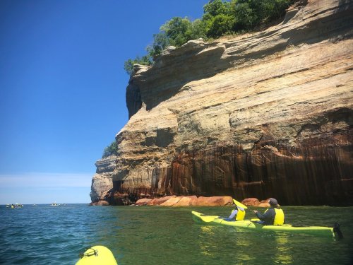 Two kayakers in yellow kayaks paddle near a towering rock cliff under a clear blue sky.