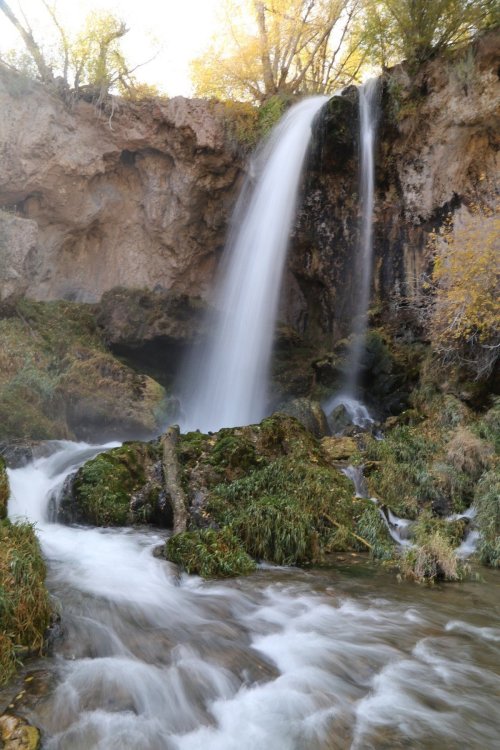 A serene waterfall cascades over rocky cliffs into a lush, green landscape with flowing water and autumn foliage.