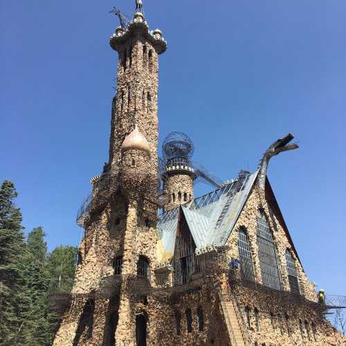 A tall, ornate stone tower with a spire, surrounded by trees and a clear blue sky.