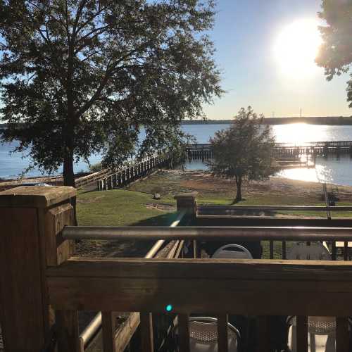 A serene lakeside view at sunset, featuring a wooden dock, trees, and calm water reflecting the sun.