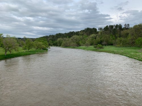 A calm river flows through a green landscape, surrounded by trees under a cloudy sky.
