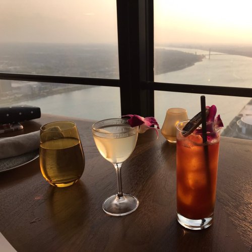 Two cocktails on a wooden table with a scenic view of a river and city skyline at sunset.