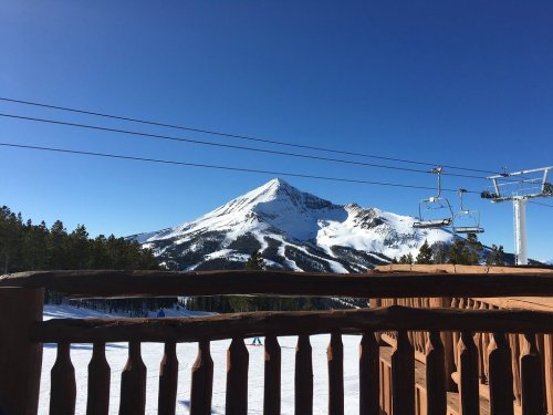 Snow-covered mountain under a clear blue sky, with ski lifts and a wooden railing in the foreground.