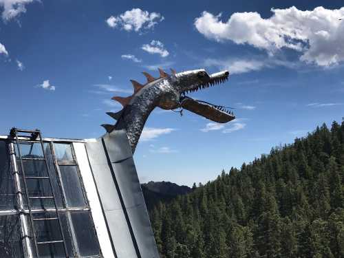 A metallic dragon sculpture perched on a rooftop, overlooking a forested mountain landscape under a blue sky.