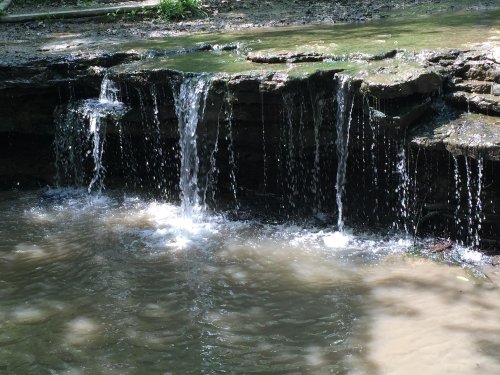 A small waterfall cascading over rocky ledges into a calm pool of water below, surrounded by greenery.