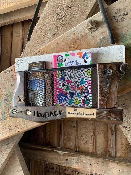 A wooden display with a mesh front holding a colorful journal, hanging on a rustic wall covered in signatures and drawings.