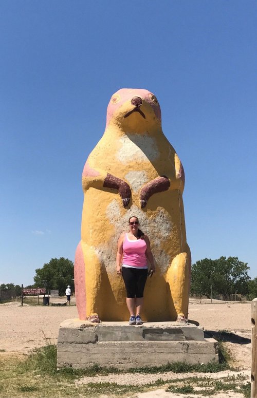 A woman stands next to a large, colorful statue of a prairie dog in a sunny outdoor setting.