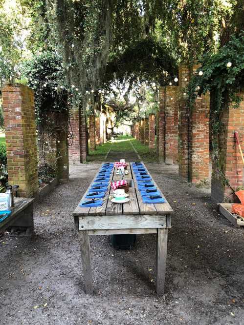 A long wooden table set with blue plates, surrounded by brick columns and greenery in a garden setting.