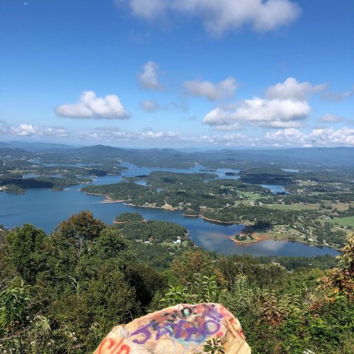 A scenic view from a mountain overlooking a lake and lush green landscape under a partly cloudy sky.
