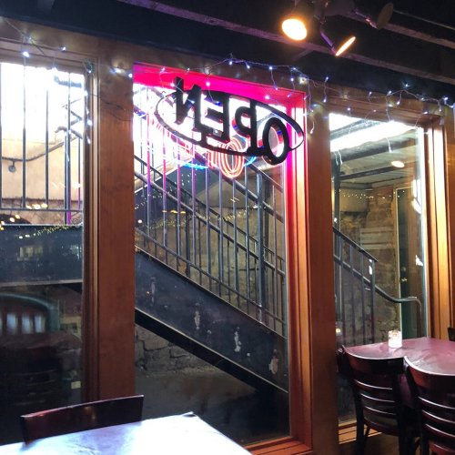 A dimly lit bar interior with an "OPEN" sign and a view of stairs through a large window.