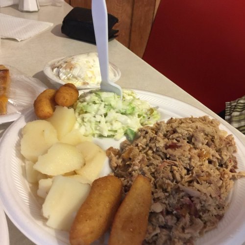 A plate of shredded meat, potato slices, coleslaw, and fried cornbread, with a dessert in the background.