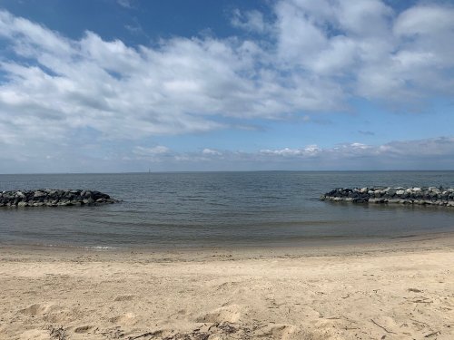 A serene beach scene with calm water, sandy shore, and rocky jetties under a partly cloudy sky.