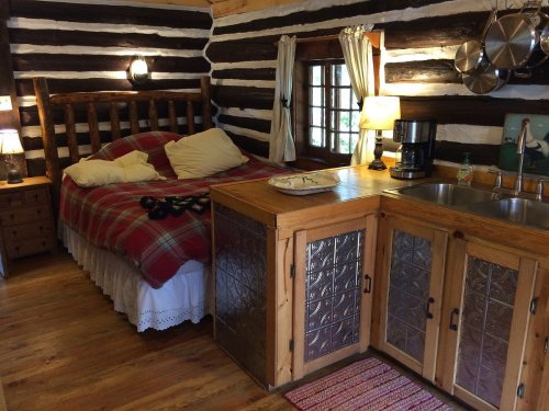 Cozy cabin interior featuring a wooden bed, kitchenette, and rustic decor with logs and warm lighting.