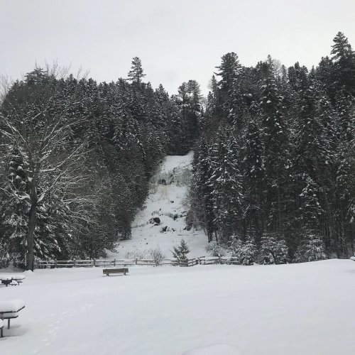 A snowy landscape with a waterfall cascading down a forested hillside, surrounded by tall evergreen trees.