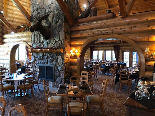 Cozy log cabin restaurant interior with wooden beams, stone fireplace, and tables set for dining.