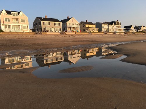 Beachfront houses reflecting in a shallow pool of water on the sand during sunset.