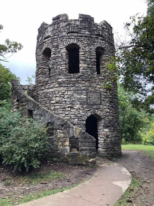 Stone tower with arched windows and a spiral staircase, surrounded by greenery and a winding path.