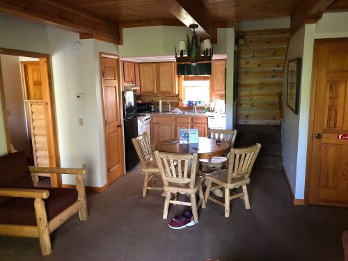 Cozy cabin interior featuring a wooden dining table, kitchen, and staircase, with rustic decor and warm lighting.