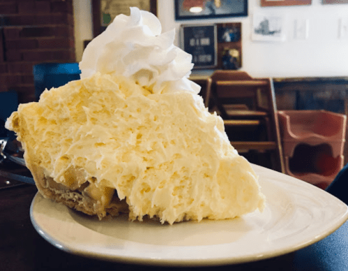 A slice of creamy pie topped with whipped cream, served on a white plate in a cozy café setting.