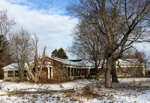 Abandoned stone building surrounded by snow and bare trees under a cloudy sky.