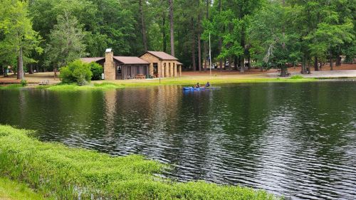 A serene lake scene with a cabin and two people kayaking, surrounded by lush green trees and grass.