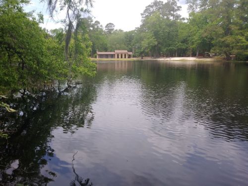 A serene lake surrounded by trees, with a small building visible on the opposite shore under a cloudy sky.