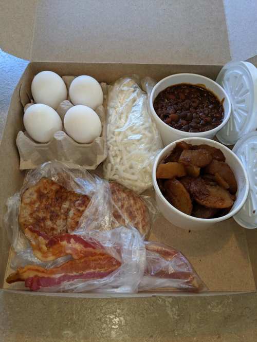 A takeout box containing eggs, shredded cheese, baked beans, apple slices, sausage patties, and bacon.