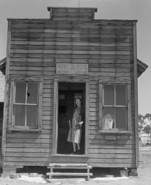 A woman stands at the entrance of a small wooden post office in Wotuse, Utah, with a sign above the door.