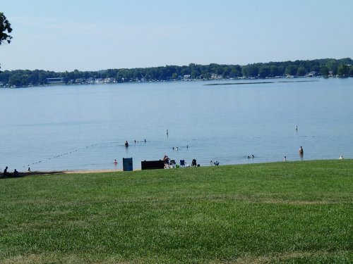 A serene lake scene with people swimming and relaxing on the beach, surrounded by green grass and trees.