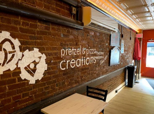 Interior of a restaurant featuring a brick wall with the text "Pretzel & Pizza Creations" and a doorway leading outside.