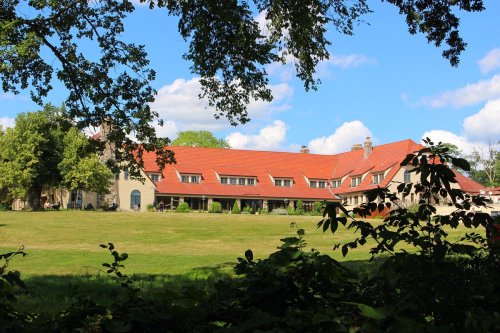 A large building with a red roof surrounded by green grass and trees under a blue sky with fluffy clouds.