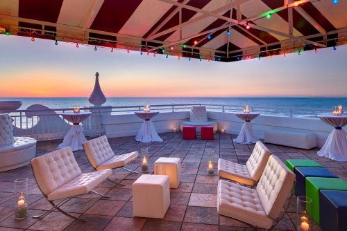 Rooftop lounge with white furniture, candles, and colorful lights overlooking a sunset by the ocean.