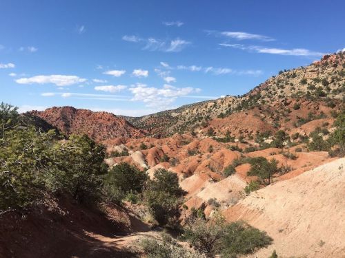 A scenic view of red rock formations and hills under a blue sky with scattered clouds. Lush greenery in the foreground.