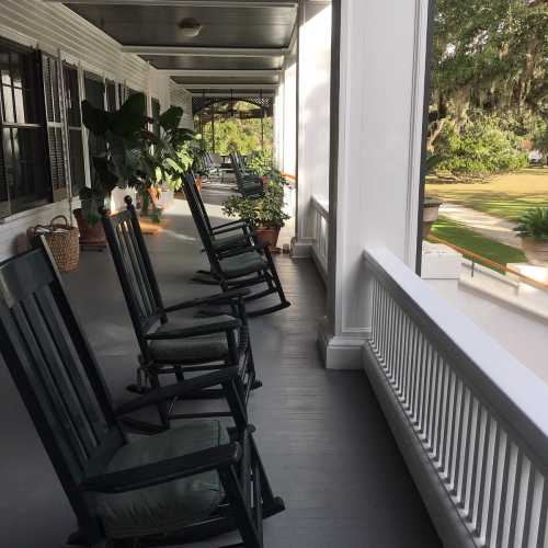 A porch with rocking chairs and potted plants, surrounded by greenery and natural light.