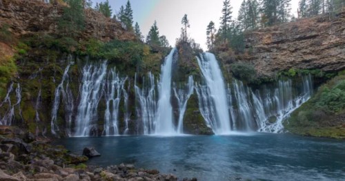 A scenic waterfall cascading over rocky cliffs into a serene blue pool, surrounded by lush green trees.