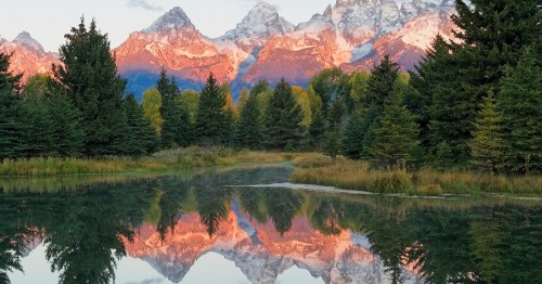A serene lake reflects majestic mountains and lush trees at sunset, creating a picturesque natural landscape.