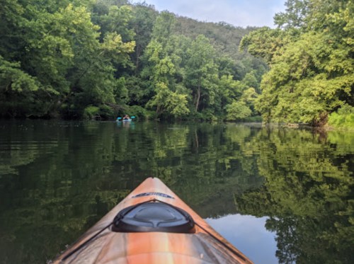 A kayak on calm water surrounded by lush green trees, with two people paddling in the distance.