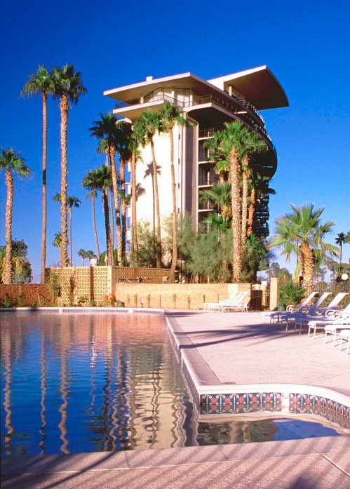 A modern building surrounded by palm trees, with a pool in the foreground reflecting the clear blue sky.