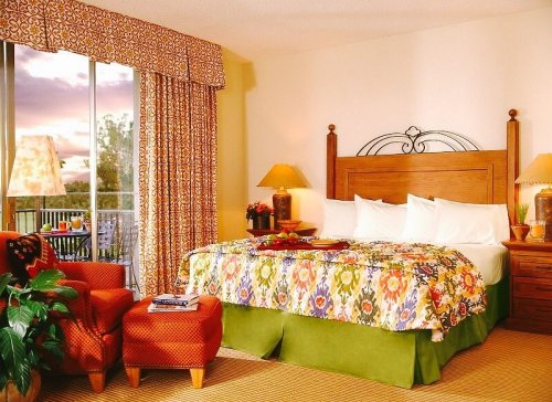 Cozy hotel room with a colorful bedspread, armchair, lamps, and large window showcasing a sunset view.