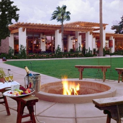 A cozy outdoor setting with a fire pit, benches, and a view of a modern building surrounded by greenery.