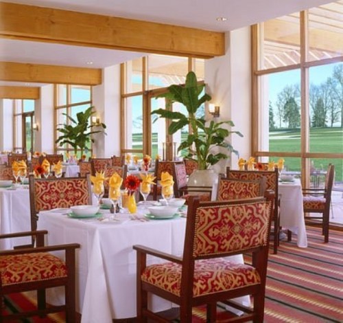 Elegant restaurant interior with wooden beams, large windows, and tables set with white linens and colorful floral arrangements.