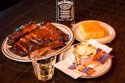 A plate of BBQ ribs with sauce, cornbread, a dessert, and a glass of whiskey on a wooden table.