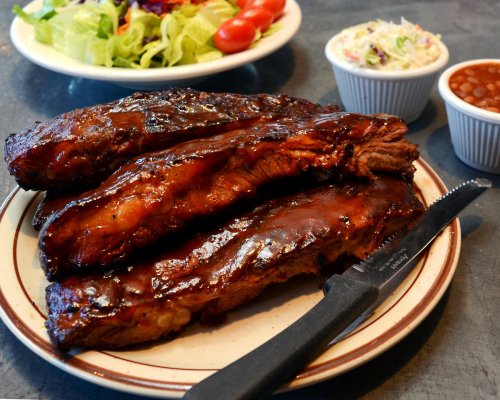 Three glazed barbecue ribs on a plate, accompanied by coleslaw, baked beans, and a fresh salad.
