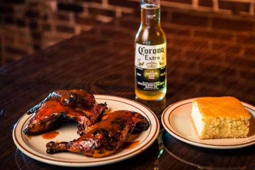 Grilled chicken with barbecue sauce, a slice of cornbread, and a bottle of Corona beer on a wooden table.