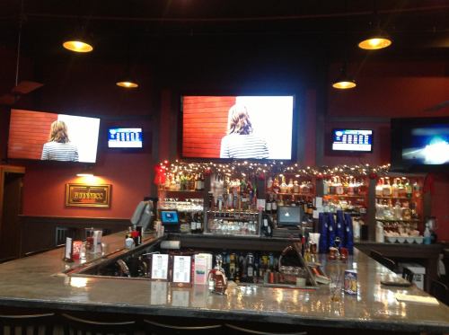 A bar interior with a long counter, shelves of liquor, and multiple TVs displaying sports or entertainment.