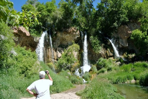 A person stands by a lush green area, photographing two cascading waterfalls surrounded by trees and grass.