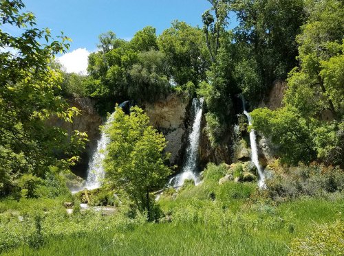 A scenic view of a waterfall cascading down rocky cliffs, surrounded by lush greenery and trees under a bright blue sky.
