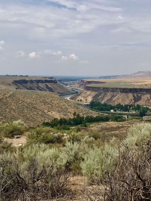 A scenic view of a winding river through a canyon, surrounded by dry hills and sparse vegetation under a cloudy sky.