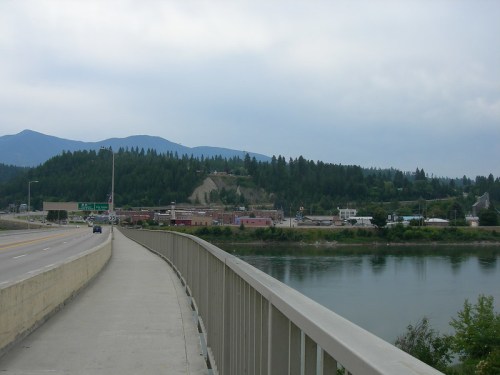 A scenic view of a river alongside a road, with mountains and trees in the background under a cloudy sky.